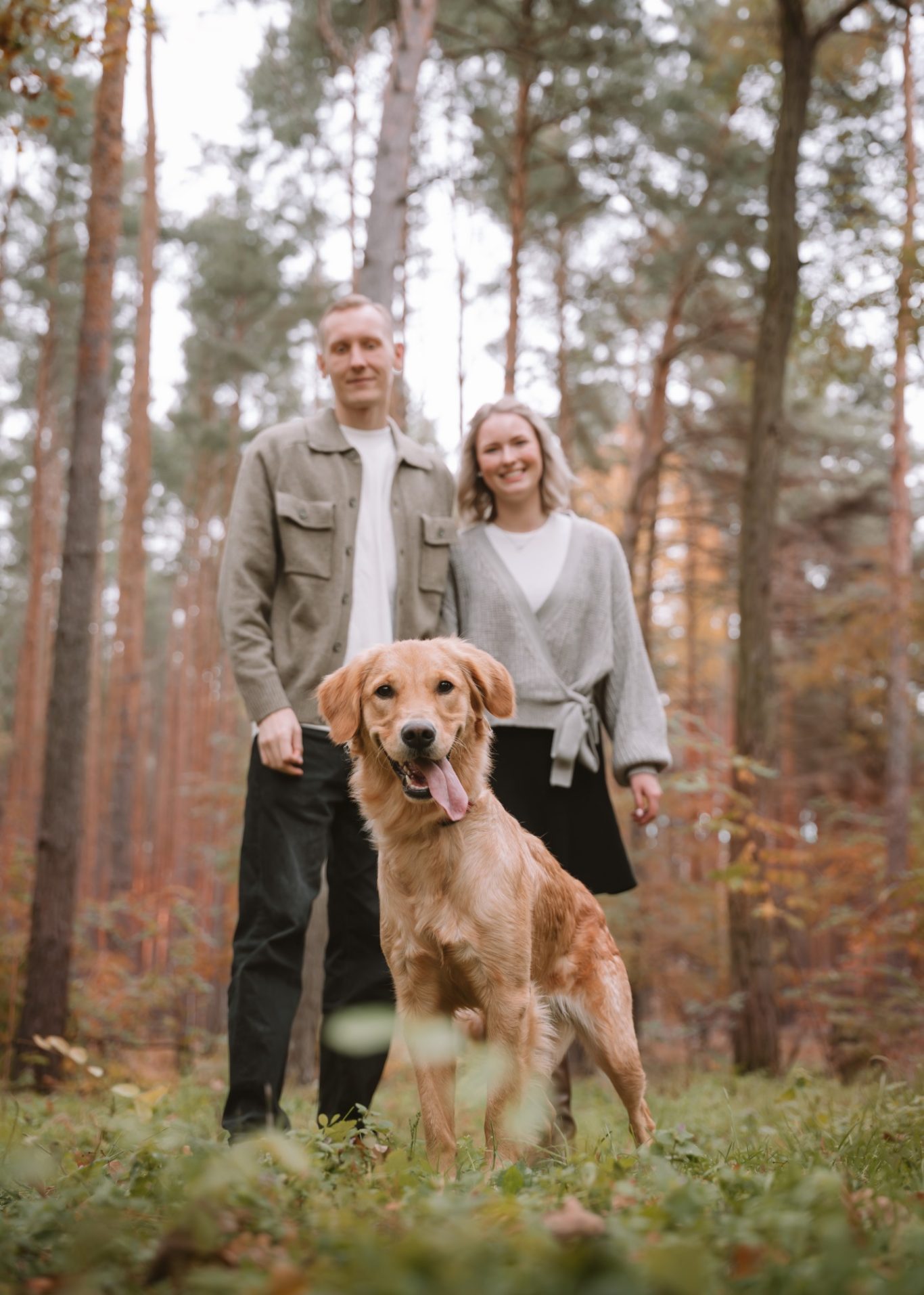 Paar steht im Wald mit einem Golden Retriever, umgeben von herbstlichen Bäumen.