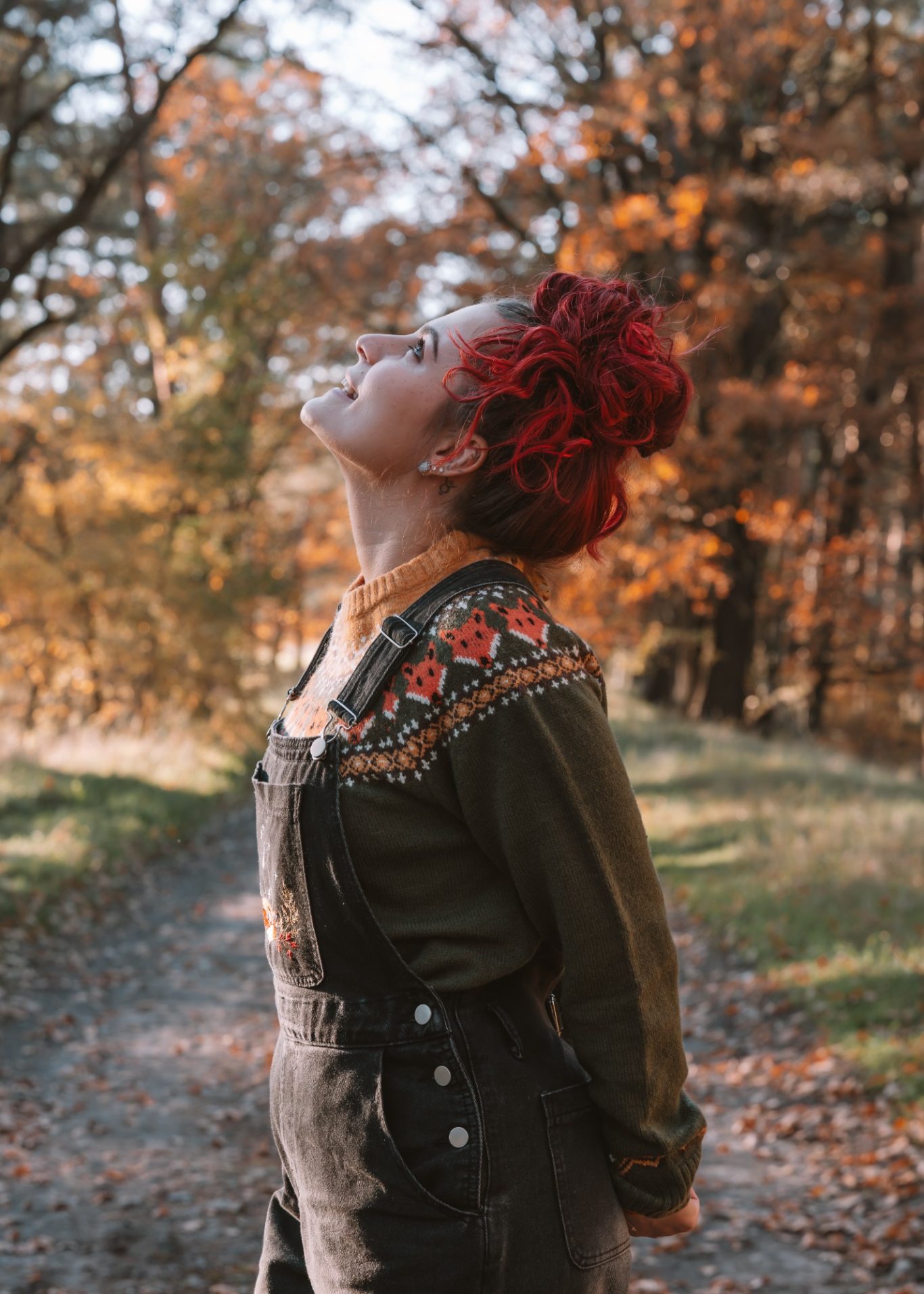 Frau mit roten Haaren und buntem Oberteil steht im herbstlichen Wald und blickt nach oben.