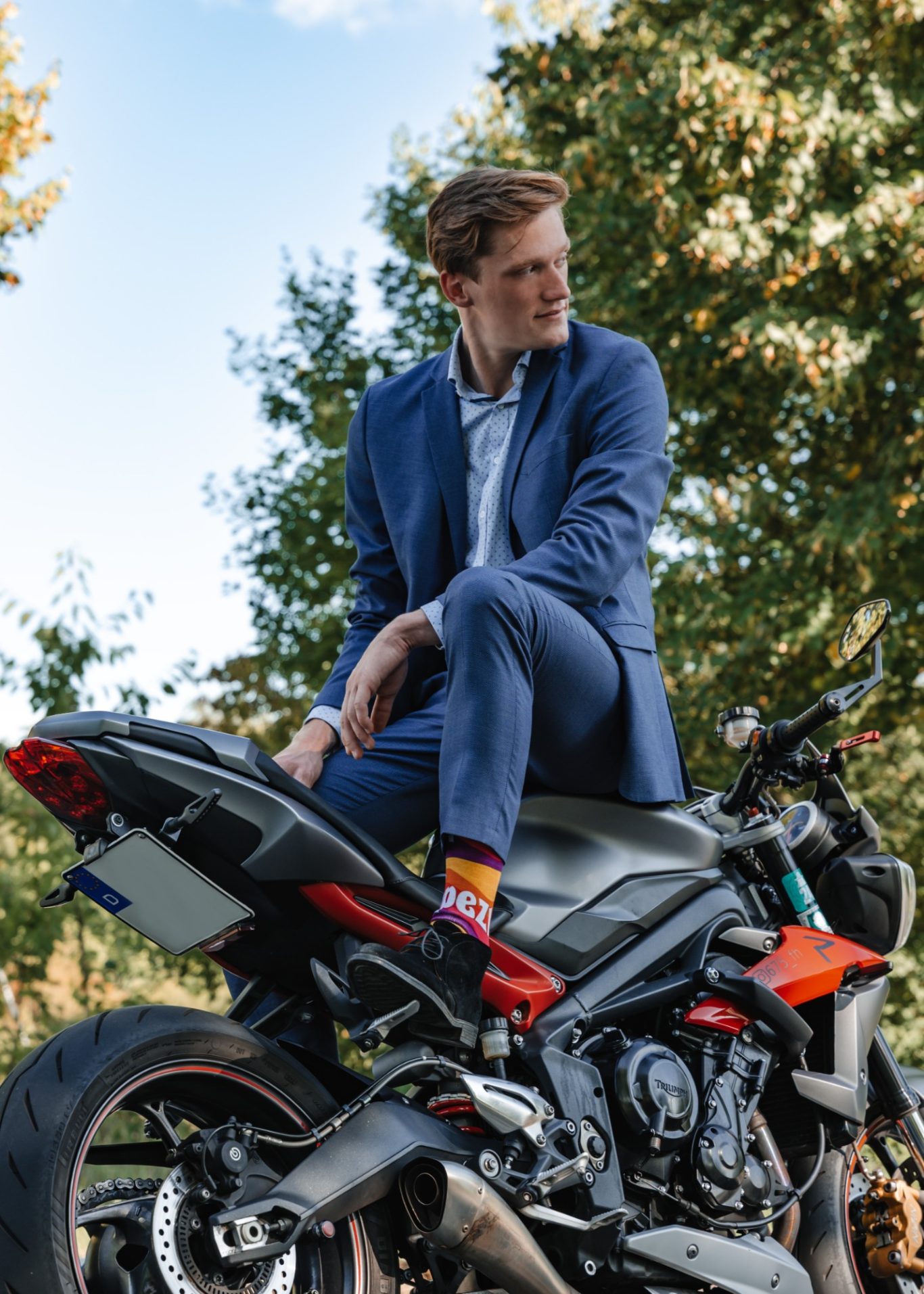 Mann im Anzug sitzt lässig auf einem Motorrad, umgeben von Bäumen.