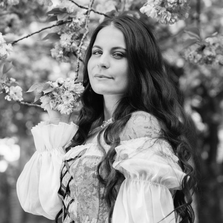 Eine junge Frau mit offenen langen, welligen und dunklen Haaren lächelt in die Kamera während sie Kirschblüten festhält.