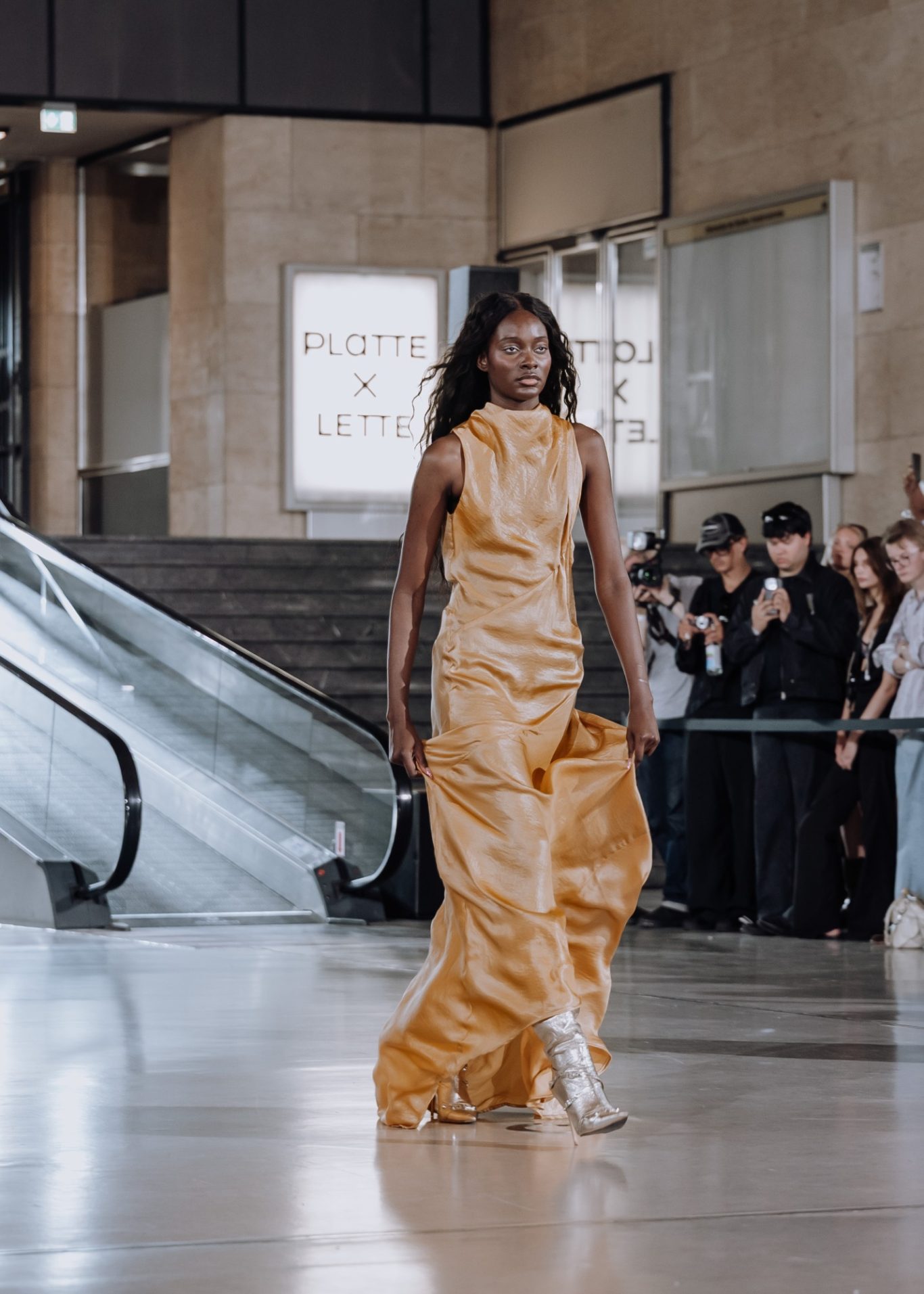 Ein Model in einem eleganten, goldenen Kleid geht an einer Rolltreppe vorbei.