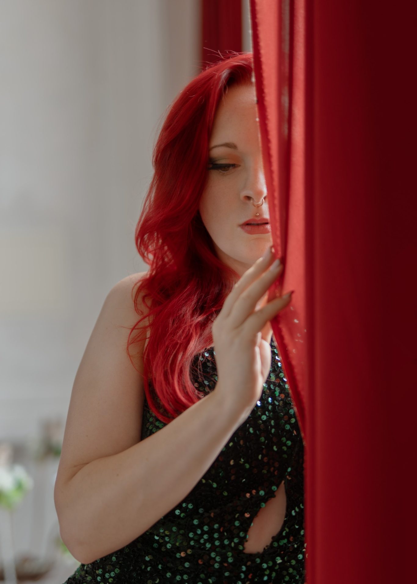 Portraitfotografie Eine junge Frau mit roten Haaren und einem glitzernden Abendkleid versteckt sich zum Teil hinter einem roten Vorhang und schaut nach unten.