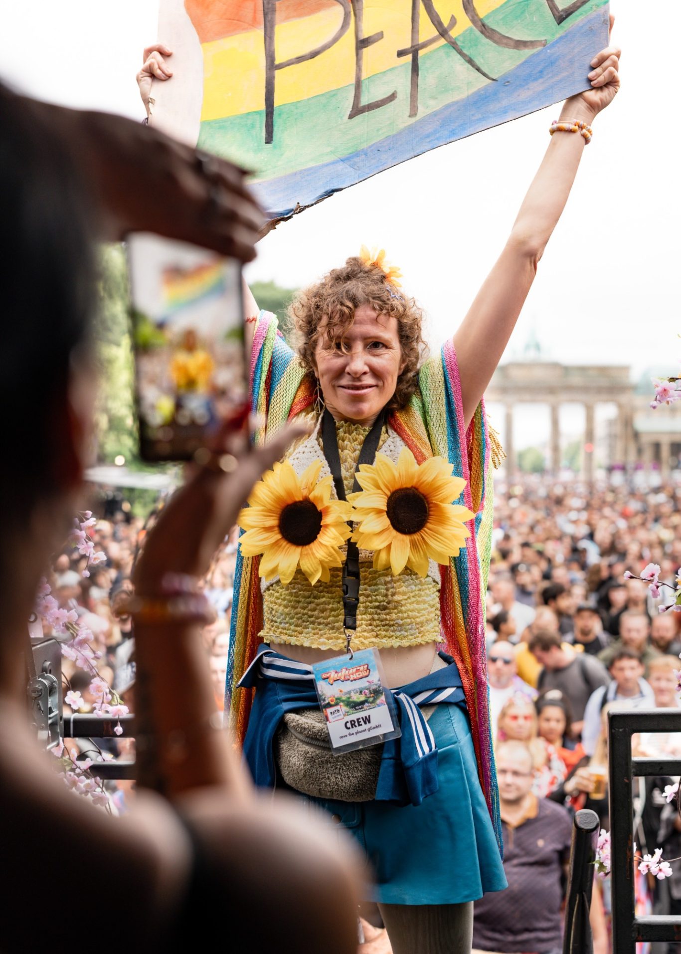 Frau mit Sonnenblumen und Regenbogenumhang hält ein "Peace"-Schild auf einer Parade.