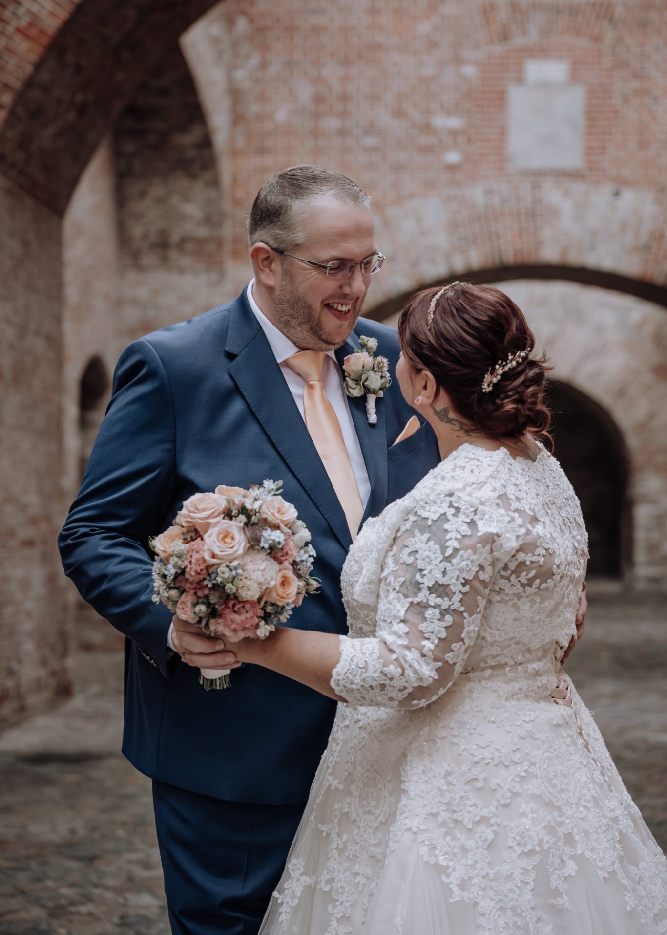 Ein Brautpaar steht sich gegenüber, lächelt und hält einen Blumenstrauß in der Hand.