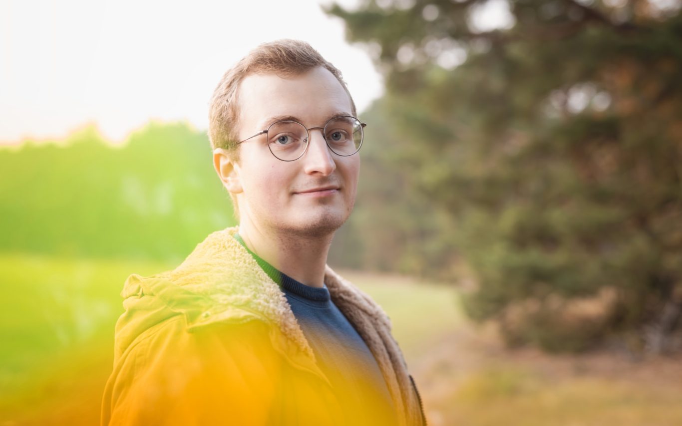 Mann mit Brille und Jacke steht in einer sonnigen Waldlandschaft.