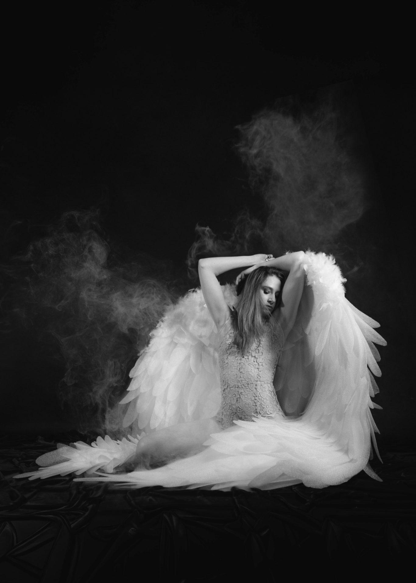 Frau mit Engelsflügeln sitzt in einer dramatischen Pose vor schwarzem Hintergrund.